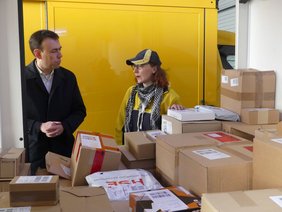 Blick durch ein Zustellfahrzeug der Post mit vielen Paketen; dahinter Nils Schmid und eine Zustellerin.