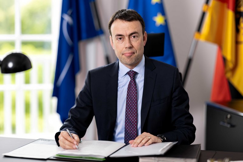 Staatssekretär Nils Schmid sitzt am Schreibtisch im Verteidigungsministerium. In der Hand hält er einen Stift, vor ihm liegen Dokumente. Im Hintergrund sind die Fahnen der NATO, der EU und der BRD zu sehen.