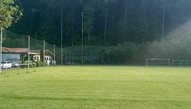 Sportplatz mit Rasen und Vereinsheim - ein Sonnenstrahl erleuchtet das Grün vor düsterem Wald.