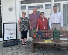 Vier Erwachsene stehen vor einem Haus. Vor ihnen steht eine bunt bemalte Bank. Neben ihnen steht ein Schild der Lebenshilfe.hinter einer bunt bemalte