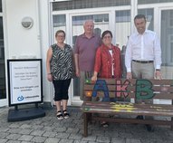 Vier Erwachsene stehen vor einem Haus. Vor ihnen steht eine bunt bemalte Bank. Neben ihnen steht ein Schild der Lebenshilfe.hinter einer bunt bemalte